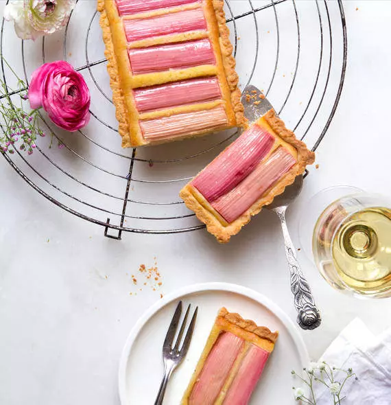 Tarte de rhubarbe présentée sur une grille avec une part découpée, entourée de fleurs et d'un verre de vin blanc.