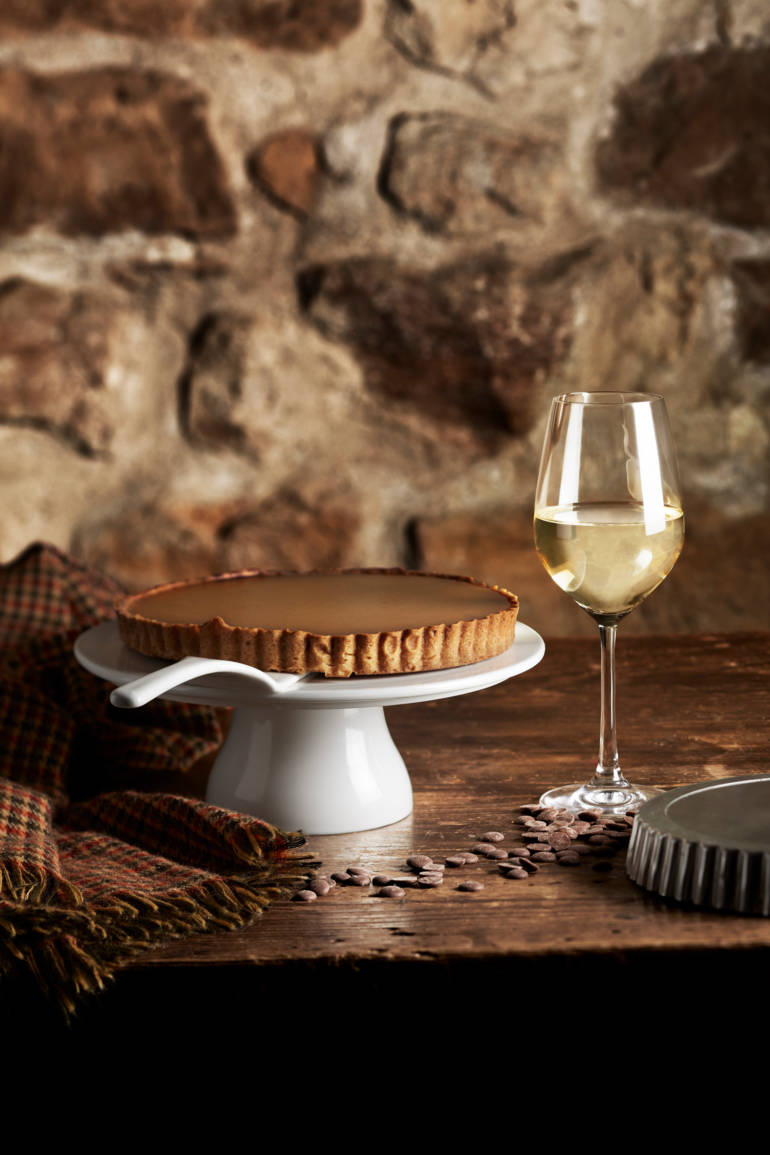 Une tarte au chocolat est présentée sur un plat blanc avec un verre de vin blanc et des fèves de cacao sur une table en bois.